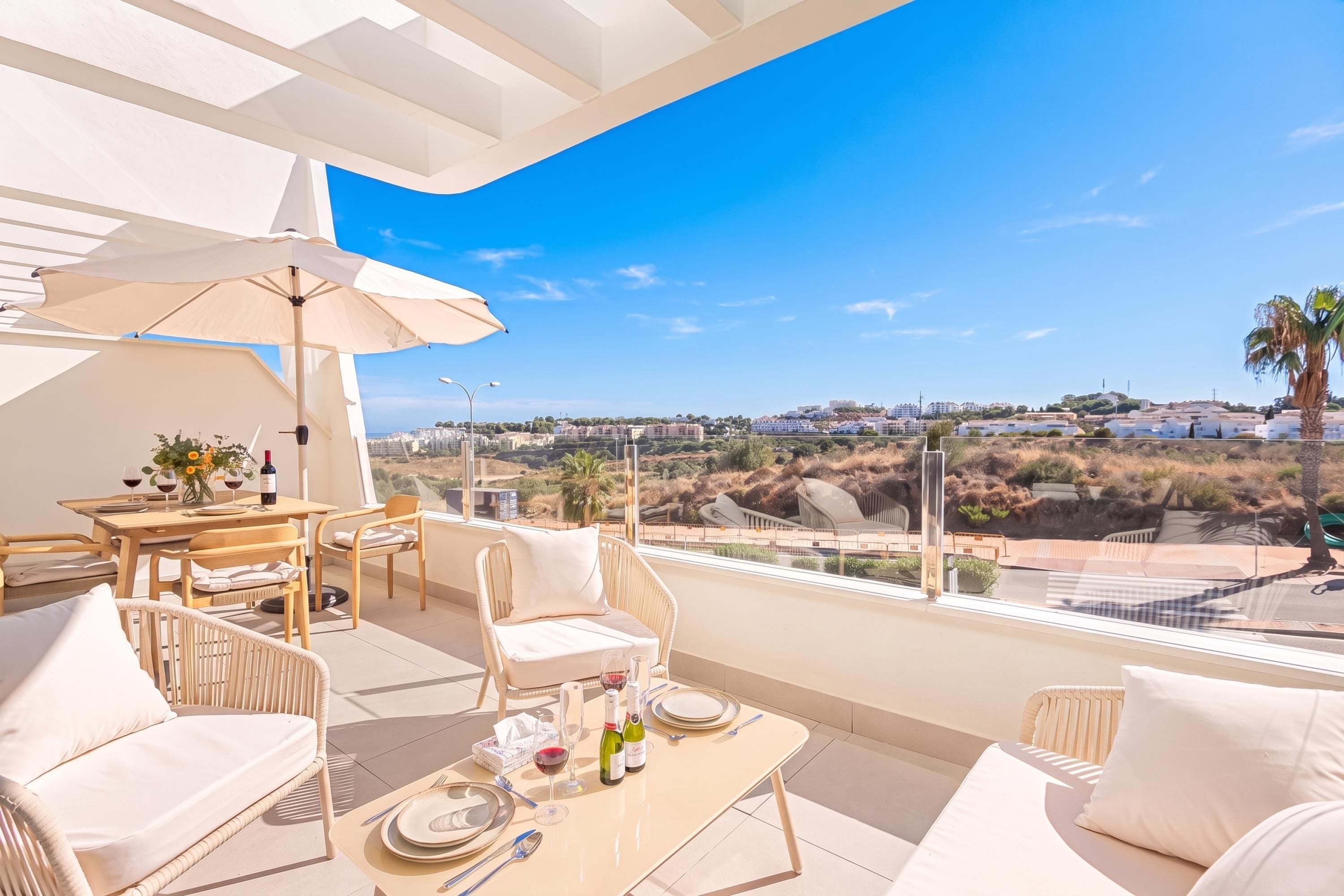 A balcony with a table set for dining, white furniture, and a view of a coastal landscape.