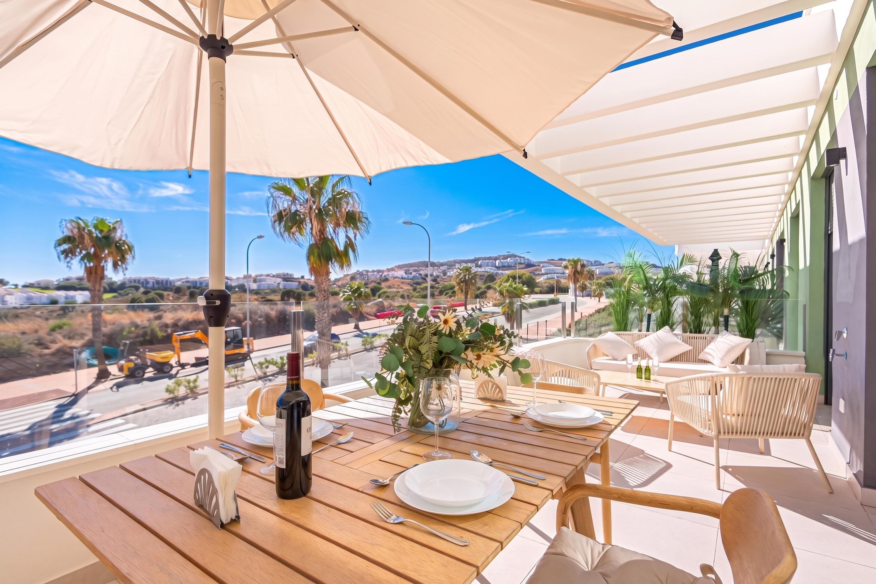 Mesa de comedor dispuesta con platos, cubiertos y una botella, bajo un paraguas grande, con vistas a un paisaje con palmeras y edificios.