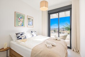 Ein Hotelzimmer mit einem Bett, einem Nachttisch, einer Lampe, zwei gerahmten Kunstwerken und Blick auf die Stadt durch ein großes Fenster.