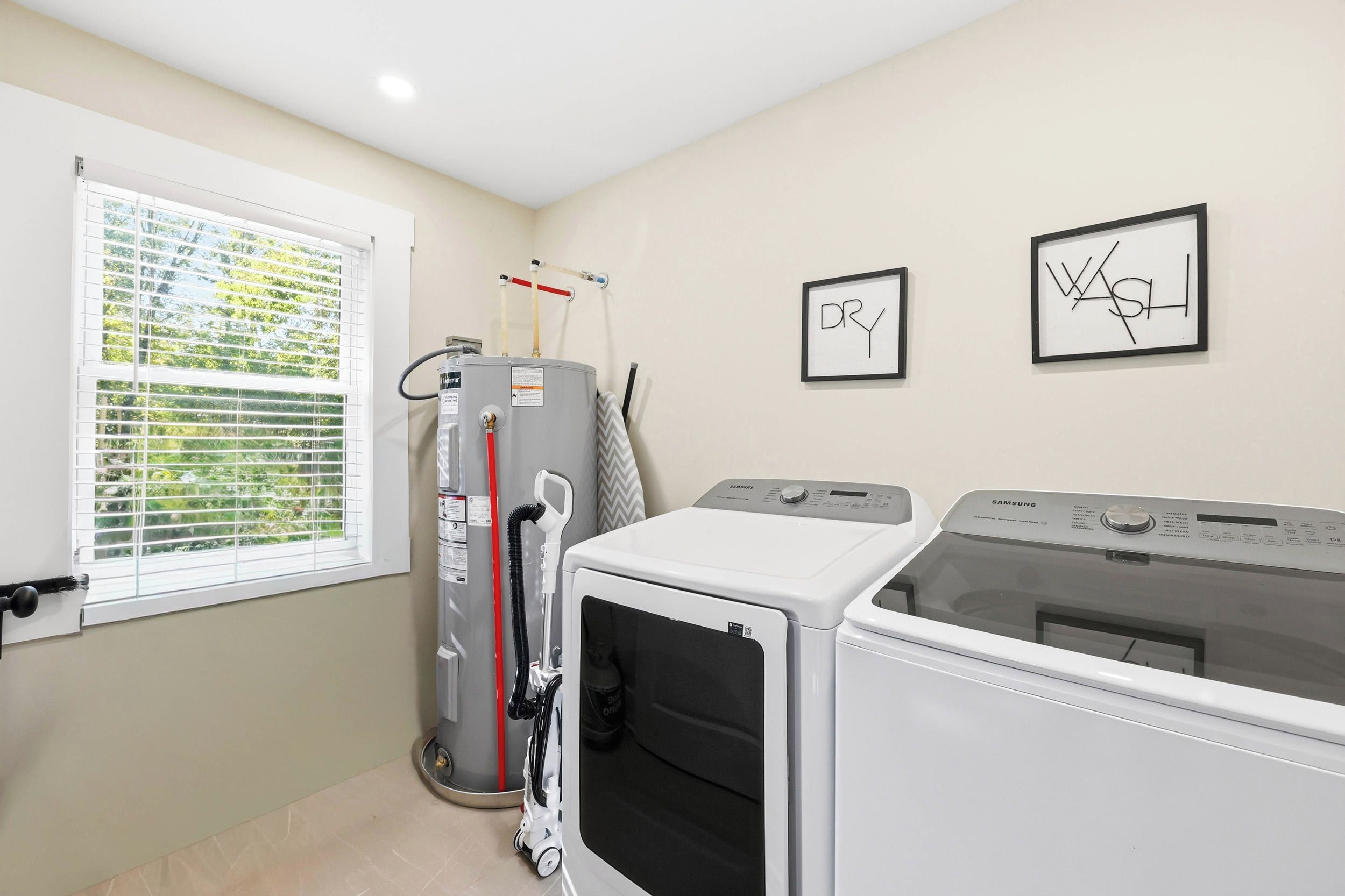 A laundry room with a large industrial washer, a smaller dryer, and a water heater. There is a window with blinds, and two framed artworks on the wall.