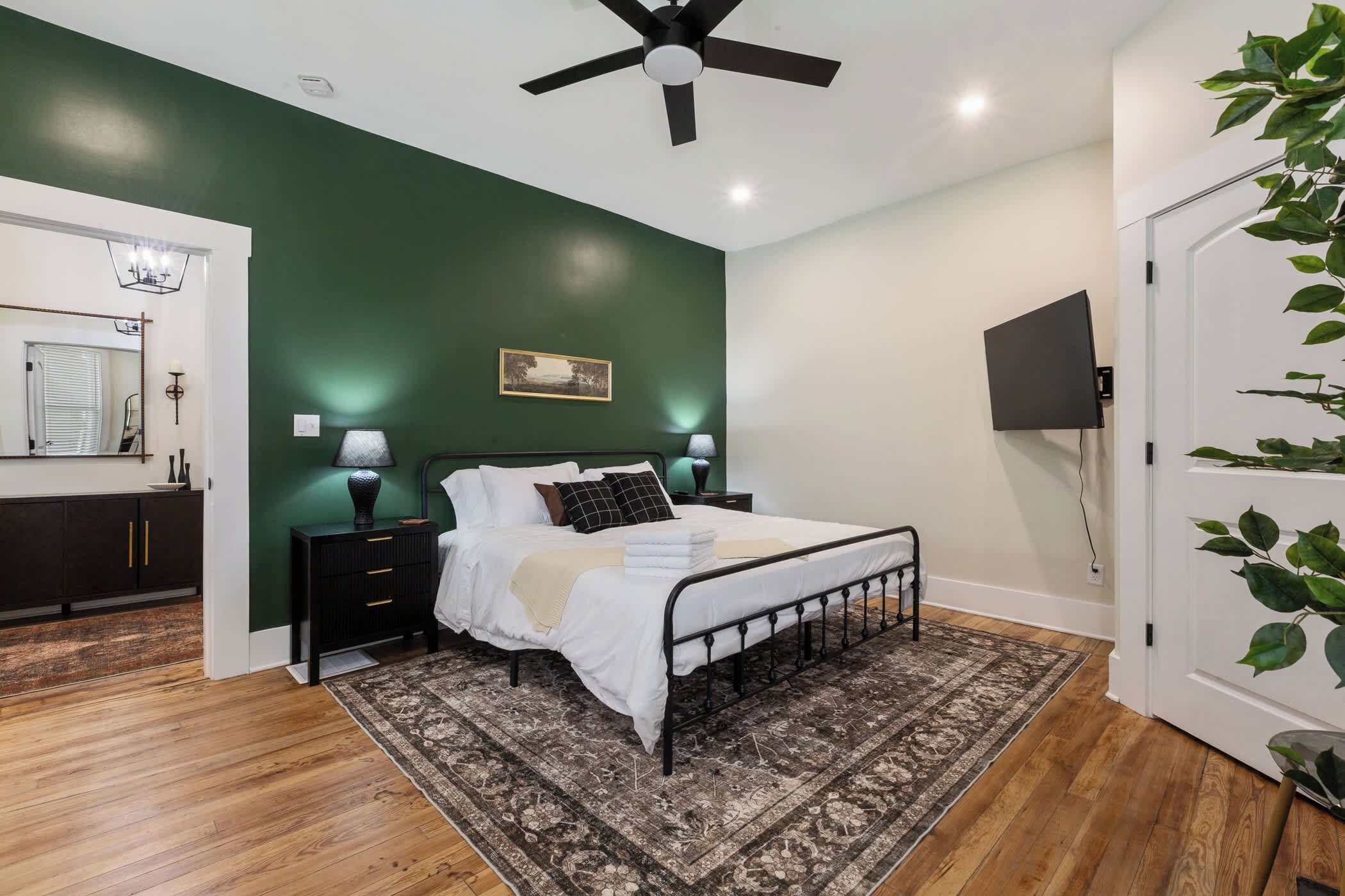 A bedroom with a bed, bedside lamps, a ceiling fan, a wooden floor, a green wall, a painting, and a television.