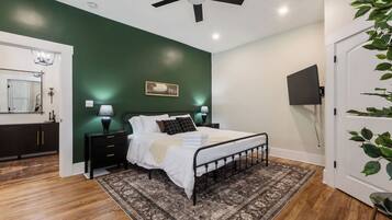 A bedroom with a bed, bedside lamps, a ceiling fan, a wooden floor, a green wall, a painting, and a television.