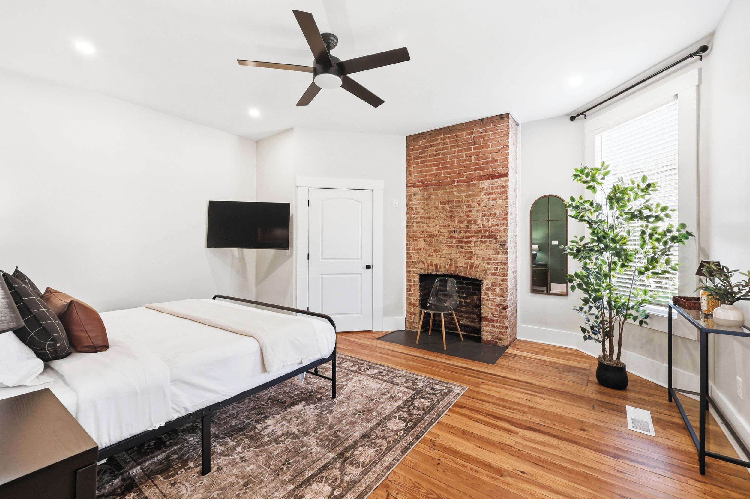 A bedroom with a bed, a ceiling fan, a brick fireplace, a TV, and a potted plant.
