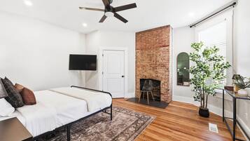A bedroom with a bed, a ceiling fan, a brick fireplace, a TV, and a potted plant.