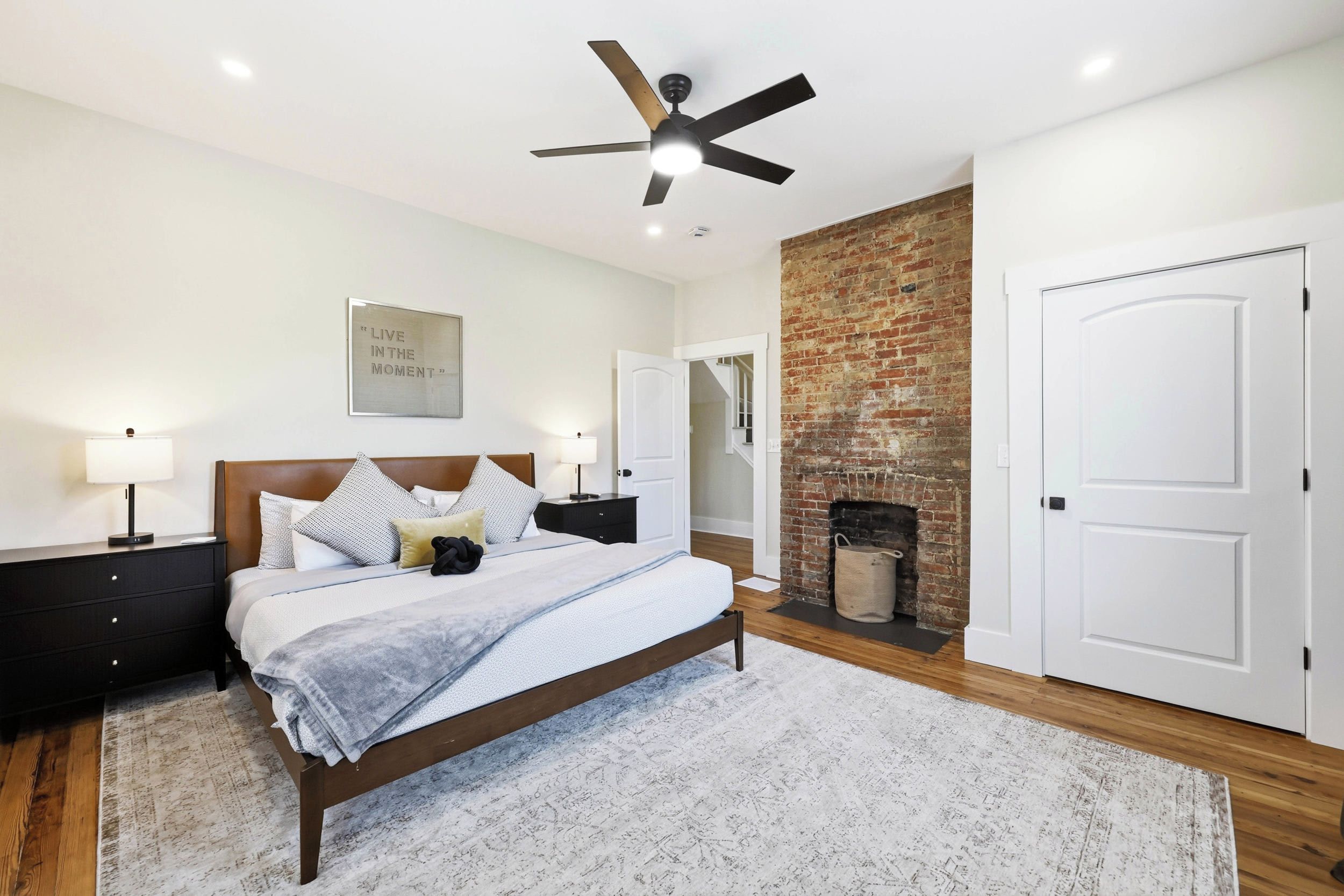 A bedroom with a bed, bedside tables, a ceiling fan, a fireplace, and a door.