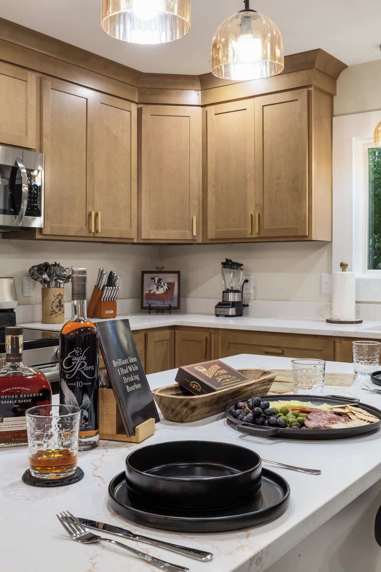 A kitchen with wooden cabinets, a microwave, and a countertop with a whiskey bottle, glasses, and a charcuterie board.