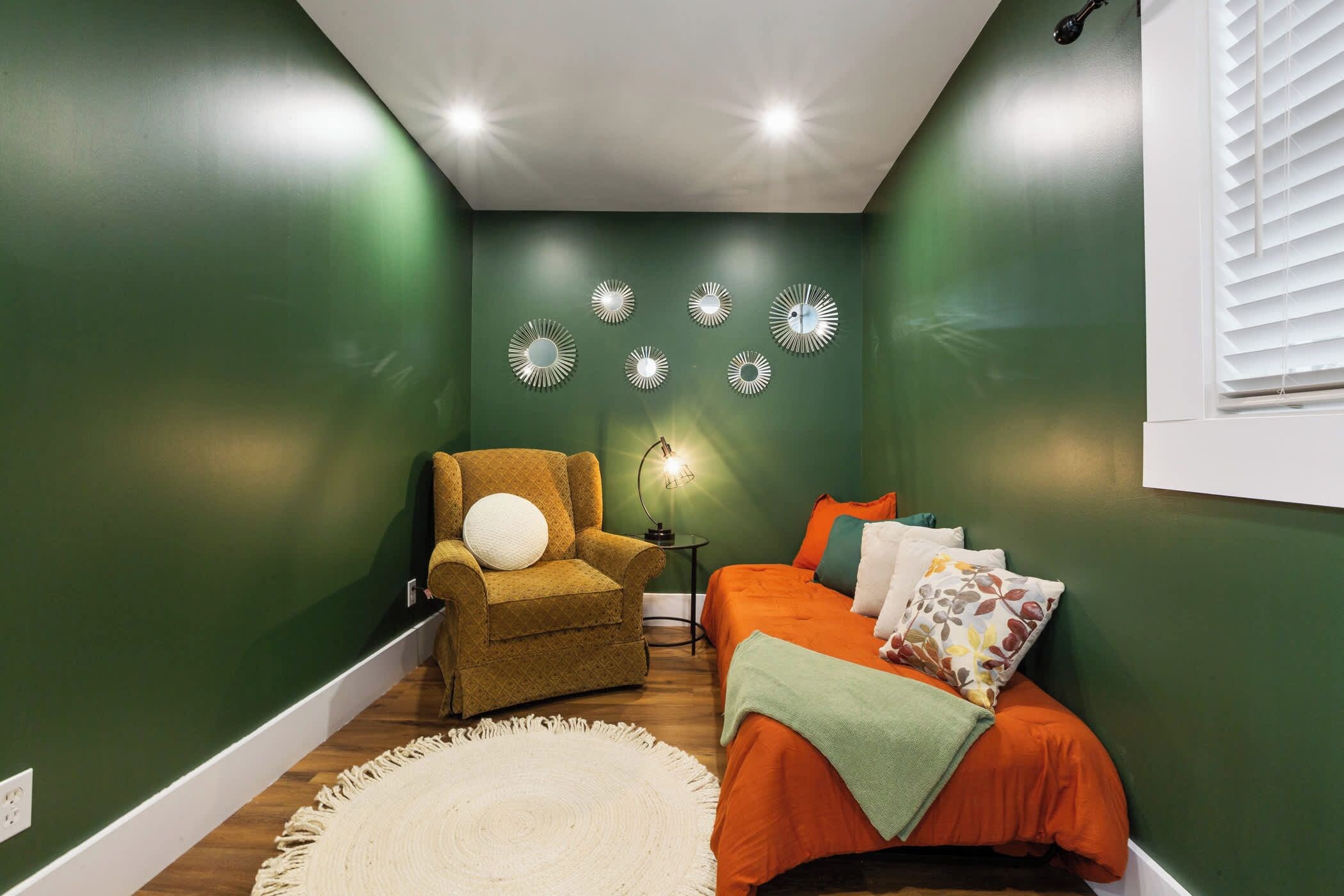 A room with green walls, a brown armchair with a white pillow, an orange bed with multiple pillows, a small table with a lamp, and decorative wall mirrors.