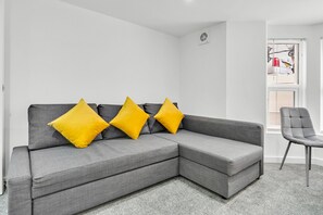 Un sofá seccional gris con tres almohadones amarillos, un sillón gris y una ventana con persianas.