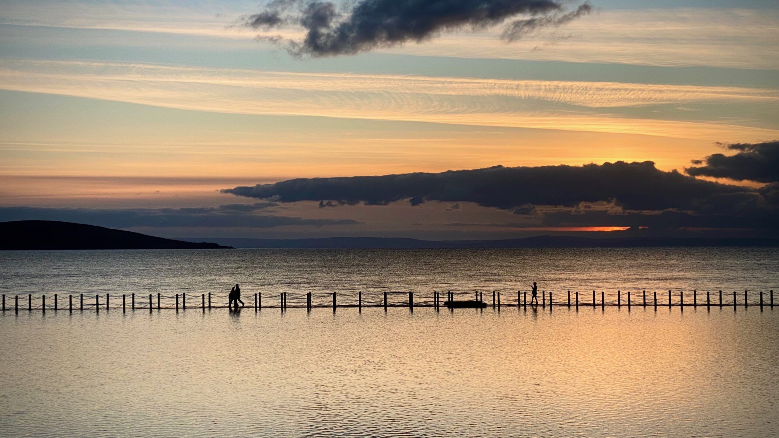 Una masa de agua tranquila con un atardecer lejano, cielo despejado y la silueta de una persona caminando por un muelle.