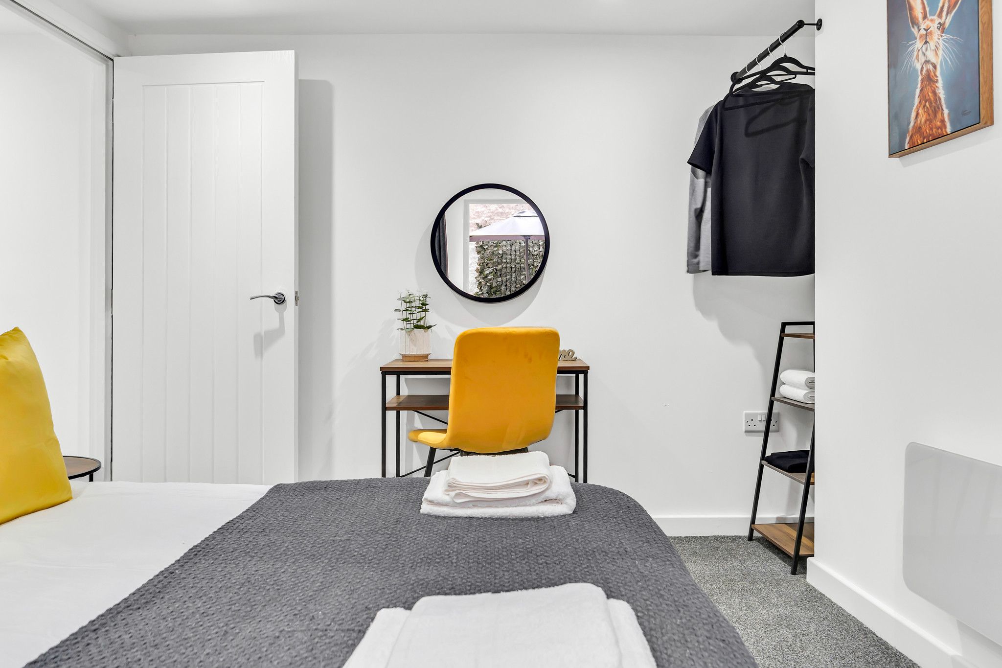A bedroom with a bed, a desk, a chair, a mirror, a hanging rack, and a painting.
