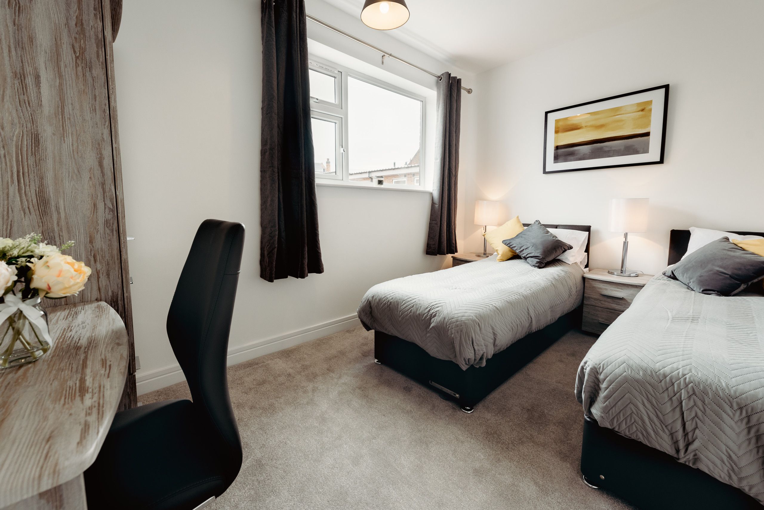 Une chambre avec deux lits, un bureau et une chaise. Il y a une fenêtre avec des rideaux, un tableau au mur et un vase rempli de fleurs sur le bureau.