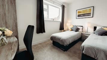 Quarto com duas camas, uma escrivaninha e uma cadeira. Há uma janela com cortinas, um quadro na parede e um vaso com flores sobre a escrivaninha.