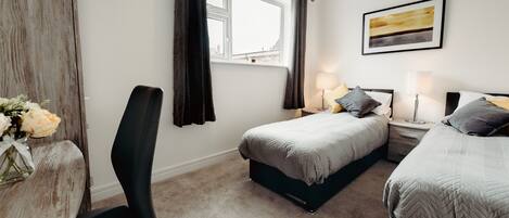 Une chambre avec deux lits, un bureau et une chaise. Il y a une fenêtre avec des rideaux, un tableau au mur et un vase rempli de fleurs sur le bureau.