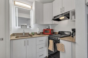 A compact kitchen with white cabinets, a stainless steel sink, a red microwave, a stove, and a small window above the sink. - Cozy Suite Near Disney + Universal (Kissimmee)