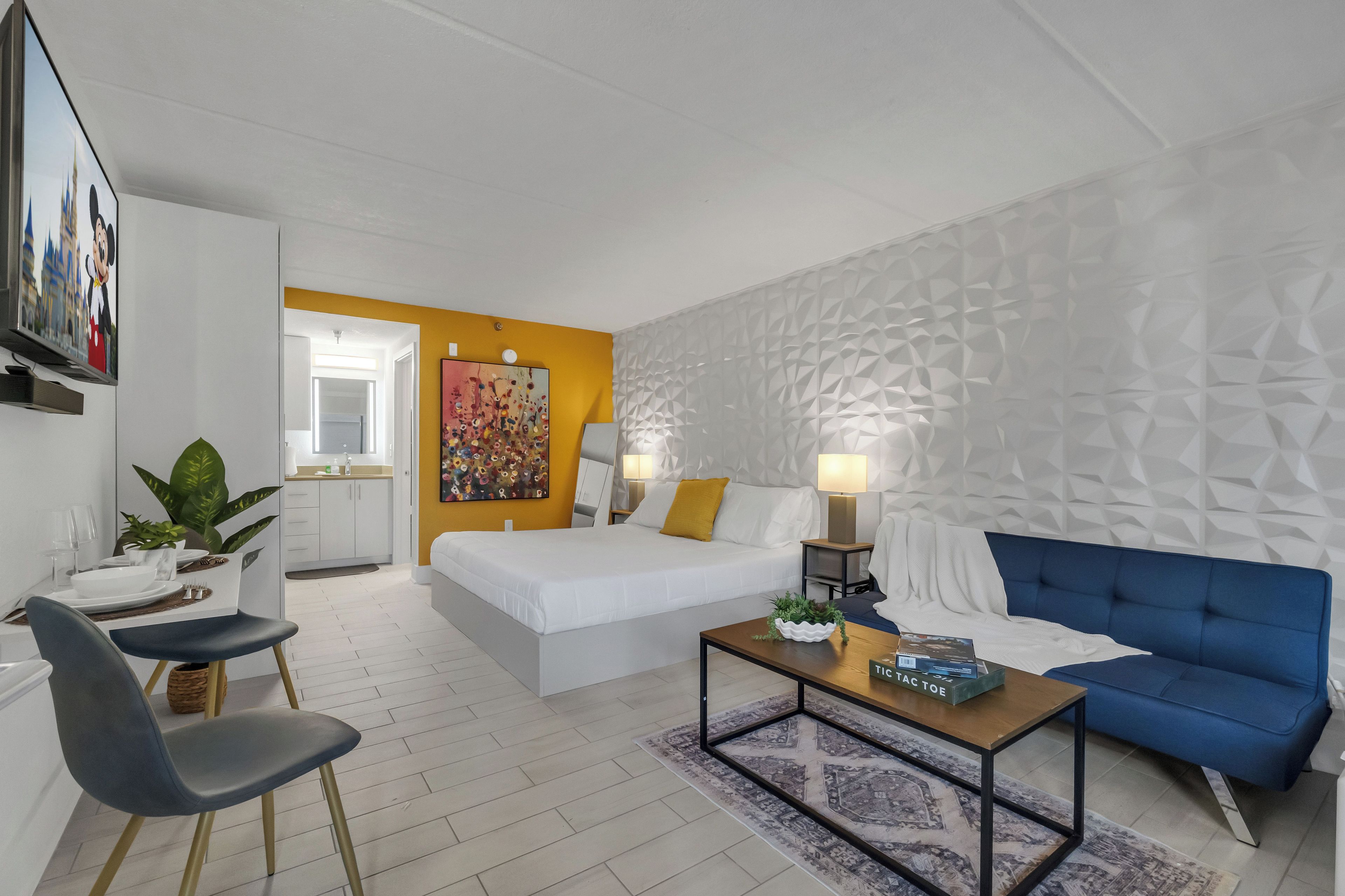 A modern hotel room with a bed, a blue sofa, a coffee table, and a dining area with chairs and a table.