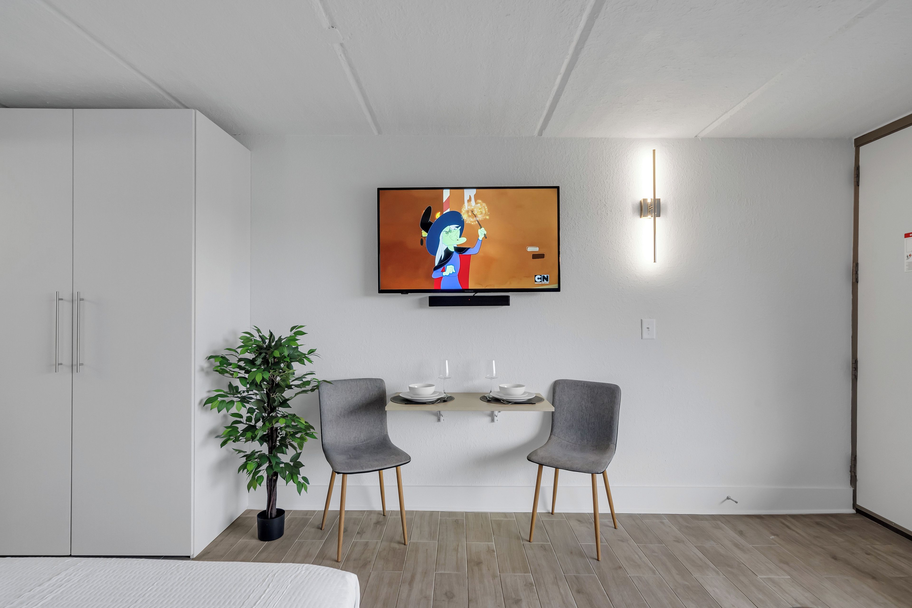 Ein modernes Zimmer mit einem an der Wand befestigten Flachbildfernseher, einem kleinen Tisch mit einer Pflanze, zwei grauen Stühlen und einem Bett mit weißer Bettdecke.