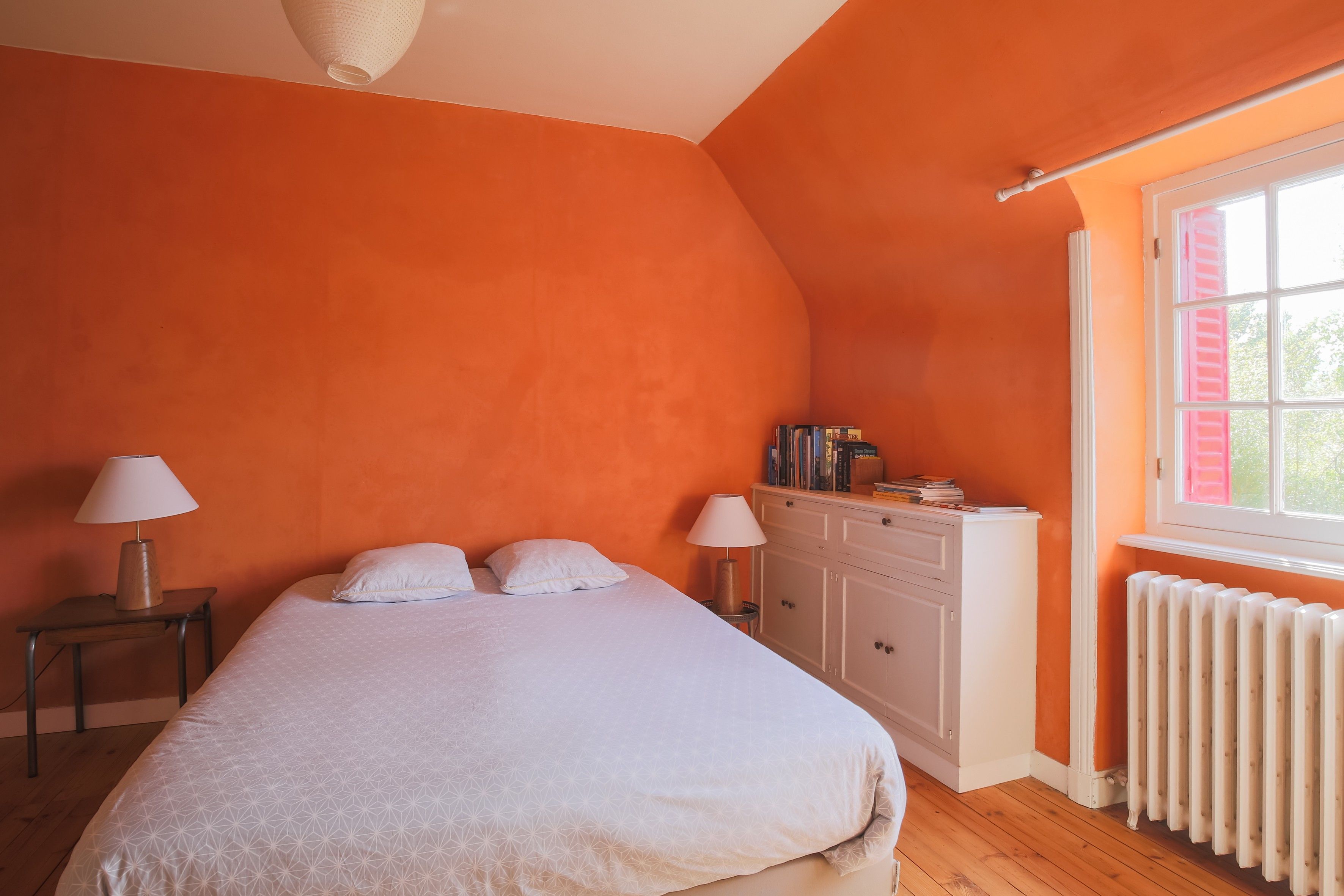 Un dormitorio con cama, dos lámparas de noche, una mesita de noche, un aparador y una ventana con contraventanas.