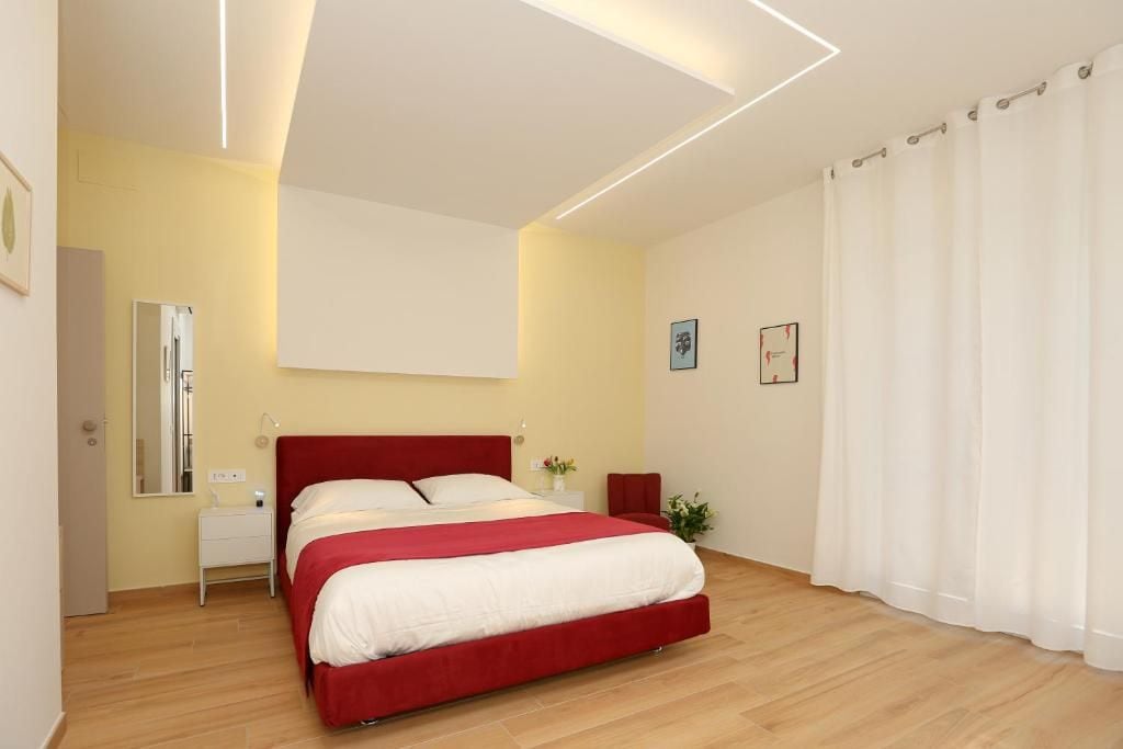 Un dormitorio moderno con una cama roja, una mesita de noche, un espejo y una ventana con cortinas.