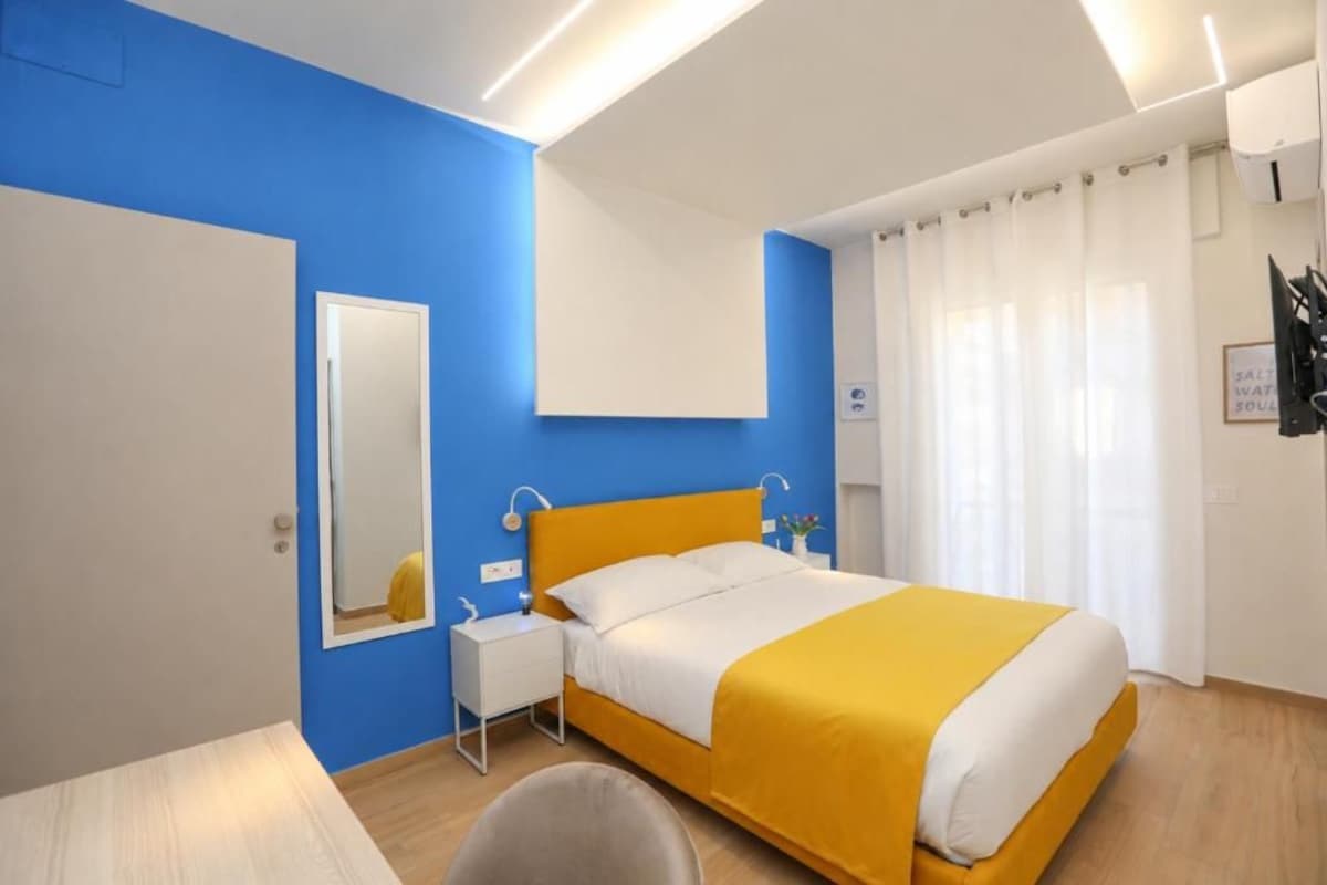 A hotel room with a bed, a bedside table, a mirror, and a window with curtains.