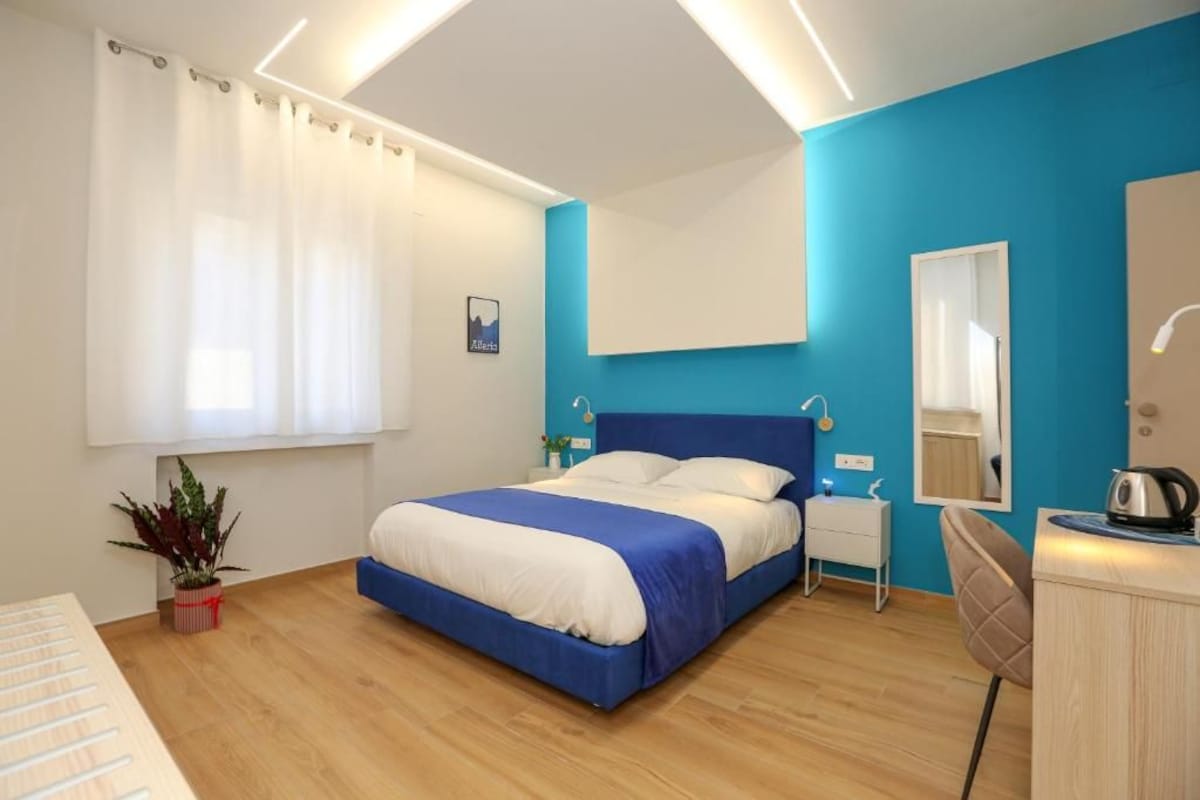 A bedroom with a bed, bedside tables, a mirror, a plant, and a desk with a kettle.