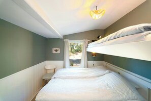 Una habitación pequeña y estrecha con una cama litera, una silla, una mesita y un ventanal con cortinas.