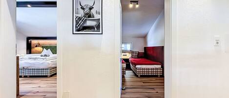 A hallway leading to two bedrooms with wooden flooring, a framed picture on the wall, and a red bedspread in one room.