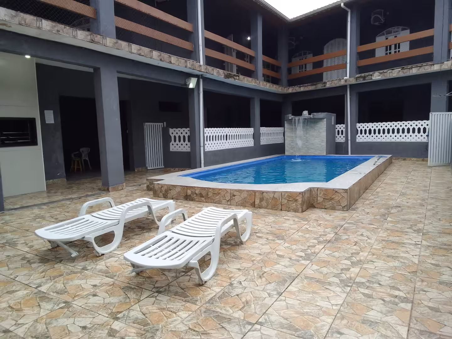A swimming pool with lounge chairs and a water feature, surrounded by a covered patio area with a tiled floor.