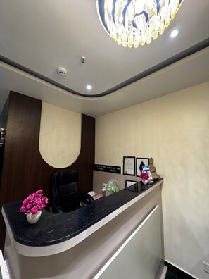 A reception desk with a black countertop, a vase of pink flowers, a seated Buddha statue, a framed certificate, and a sign indicating check-in and check-out times. - Hotel Skyrooms Inn (Varanasi)