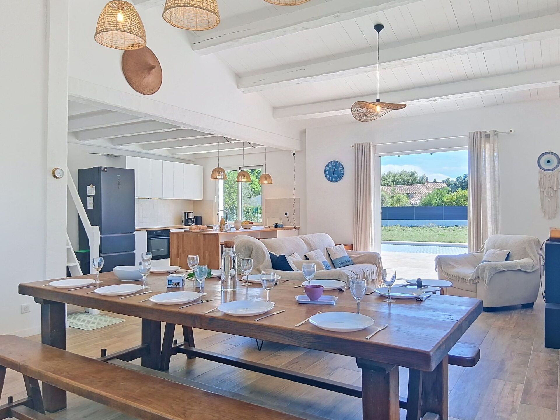 A spacious living area with a dining table set for four, a kitchen in the background, and a large window offering a view of the outdoors.