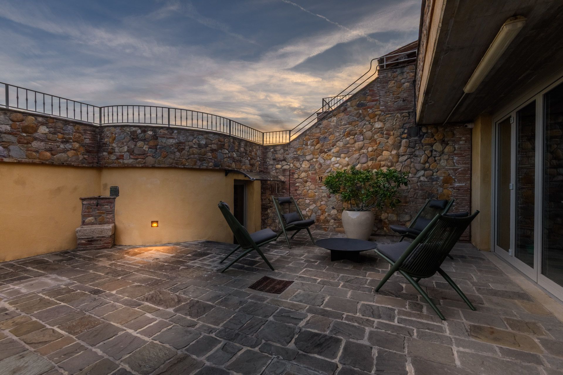 Une terrasse extérieure dotée d’un sol en pierre, d’un mur en pierre aux lignes courbes et d’un espace détente avec des chaises et une table.