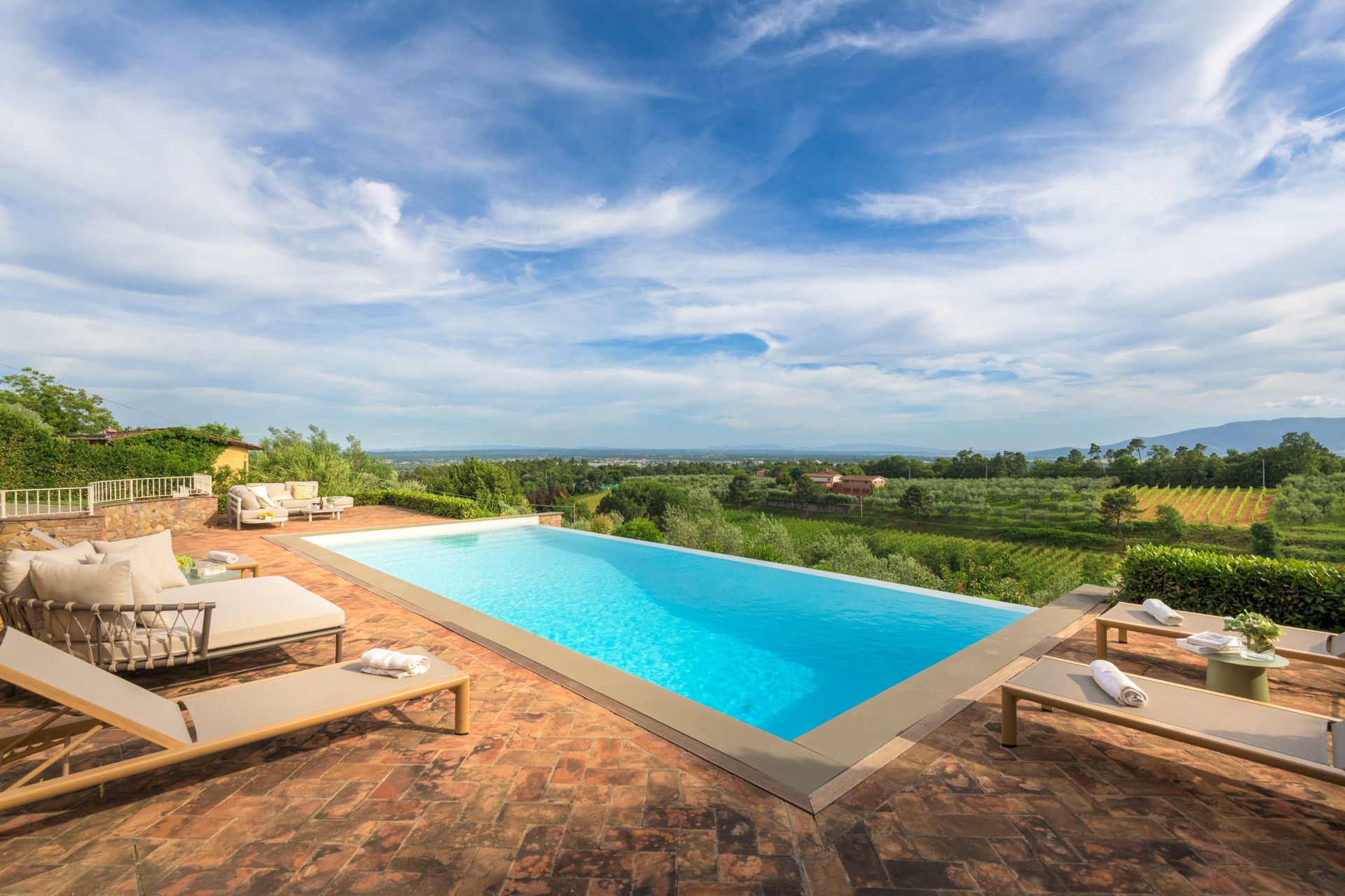 A rectangular swimming pool with a clear blue water surface, surrounded by a stone patio with lounge chairs and a table.