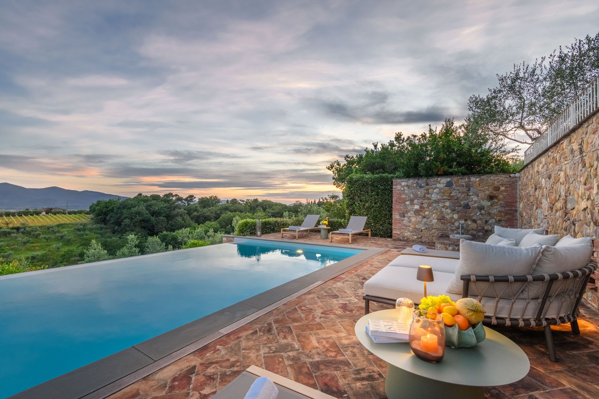 Zona junto a la piscina con pared de piedra, sillones de descanso y una mesa con libros y velas.