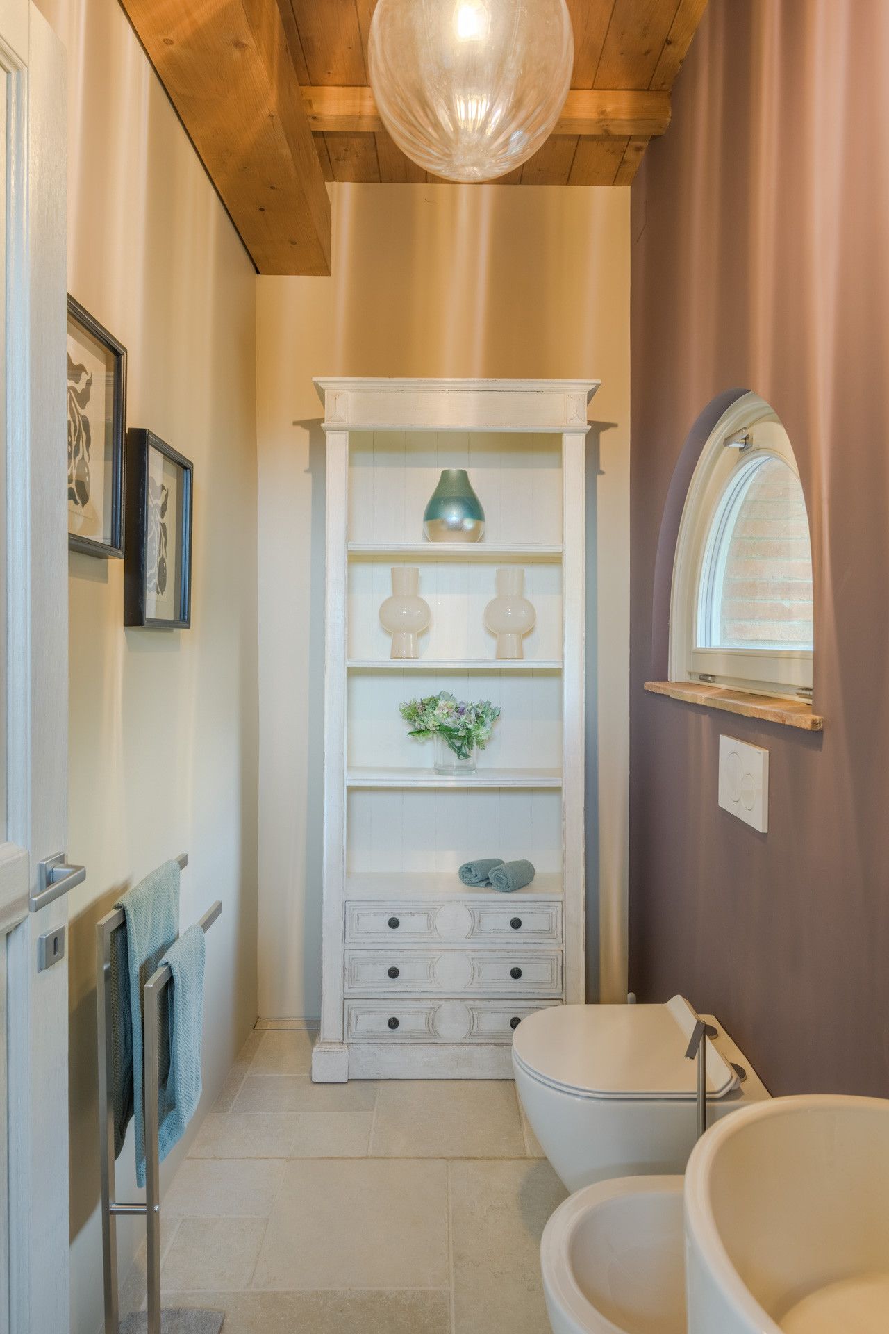 Un baño con techo de madera, un tocador blanco con espejo, un lavamanos y un inodoro. Se observa una repisa con objetos decorativos y un perchero para toallas.