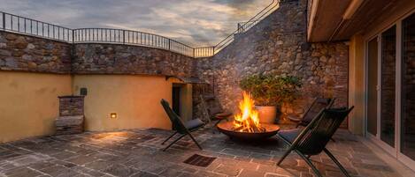 A stone patio with a fire pit and seating, surrounded by stone walls and a glass door.