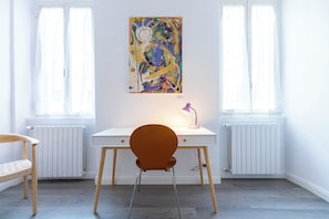 Ein Zimmer mit einem weißen Schreibtisch, einem braunen Stuhl, einem farbenfrohen abstrakten Gemälde an der Wand und einem Fenster mit weißen Vorhängen.