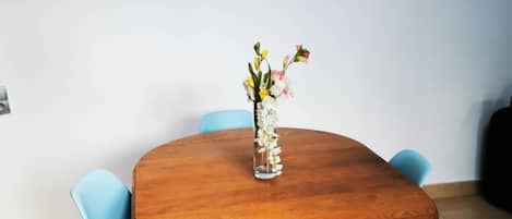 Un tavolo da pranzo rotondo in legno con un vaso di fiori, affiancato da due sedie blu, in una stanza con pavimento piastrellato e un quadro di veleggiatori al muro.