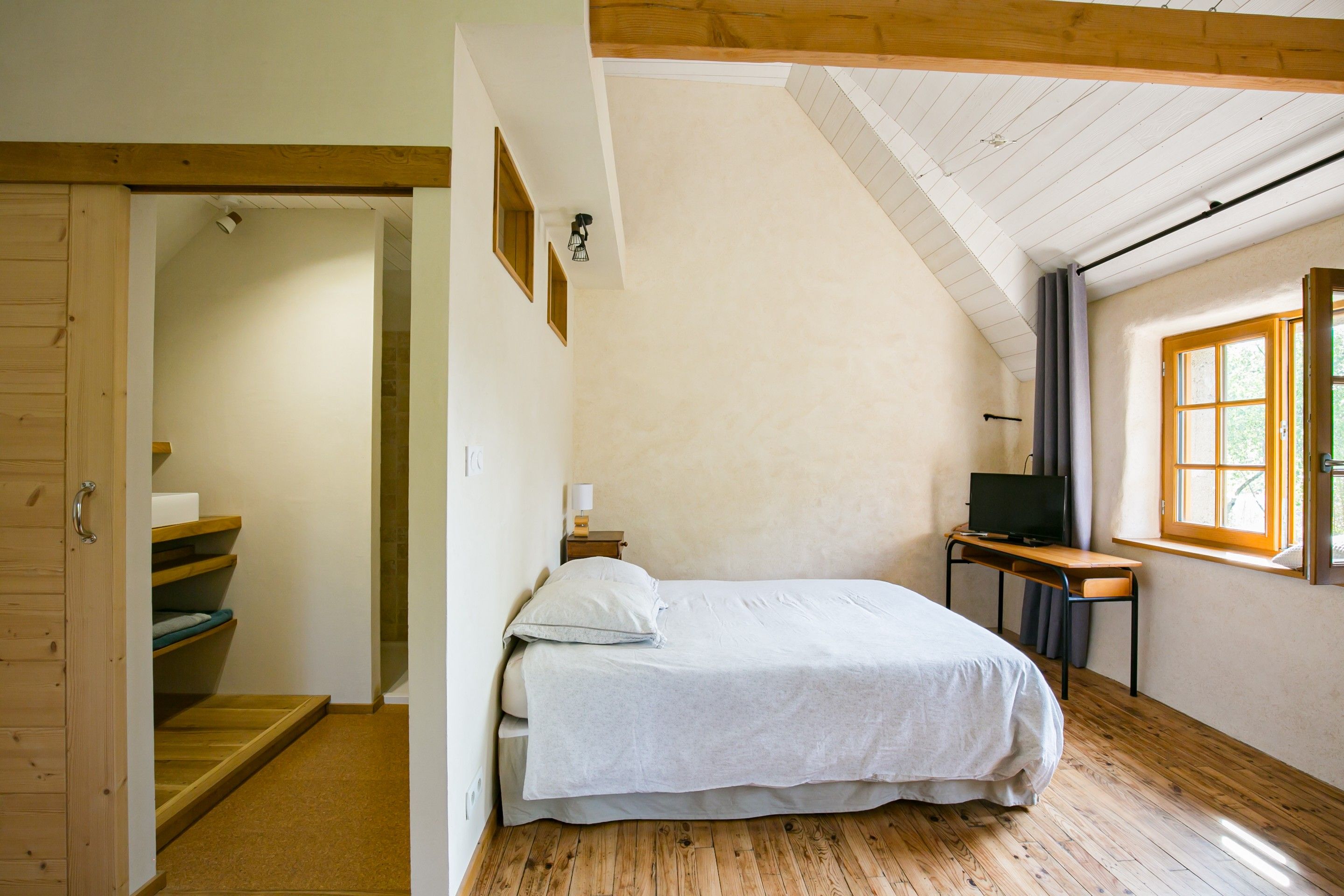 A bedroom with a bed, a desk with a computer, a window, and a wooden staircase.