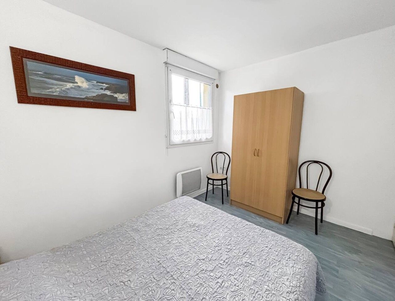 Un dormitorio con cama, armario, dos sillas, ventana con cortinas e imagen enmarcada en la pared.