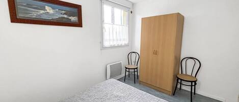 Une chambre à coucher comprenant un lit, une armoire, deux chaises, une fenêtre avec des rideaux et un tableau encadré accroché au mur.