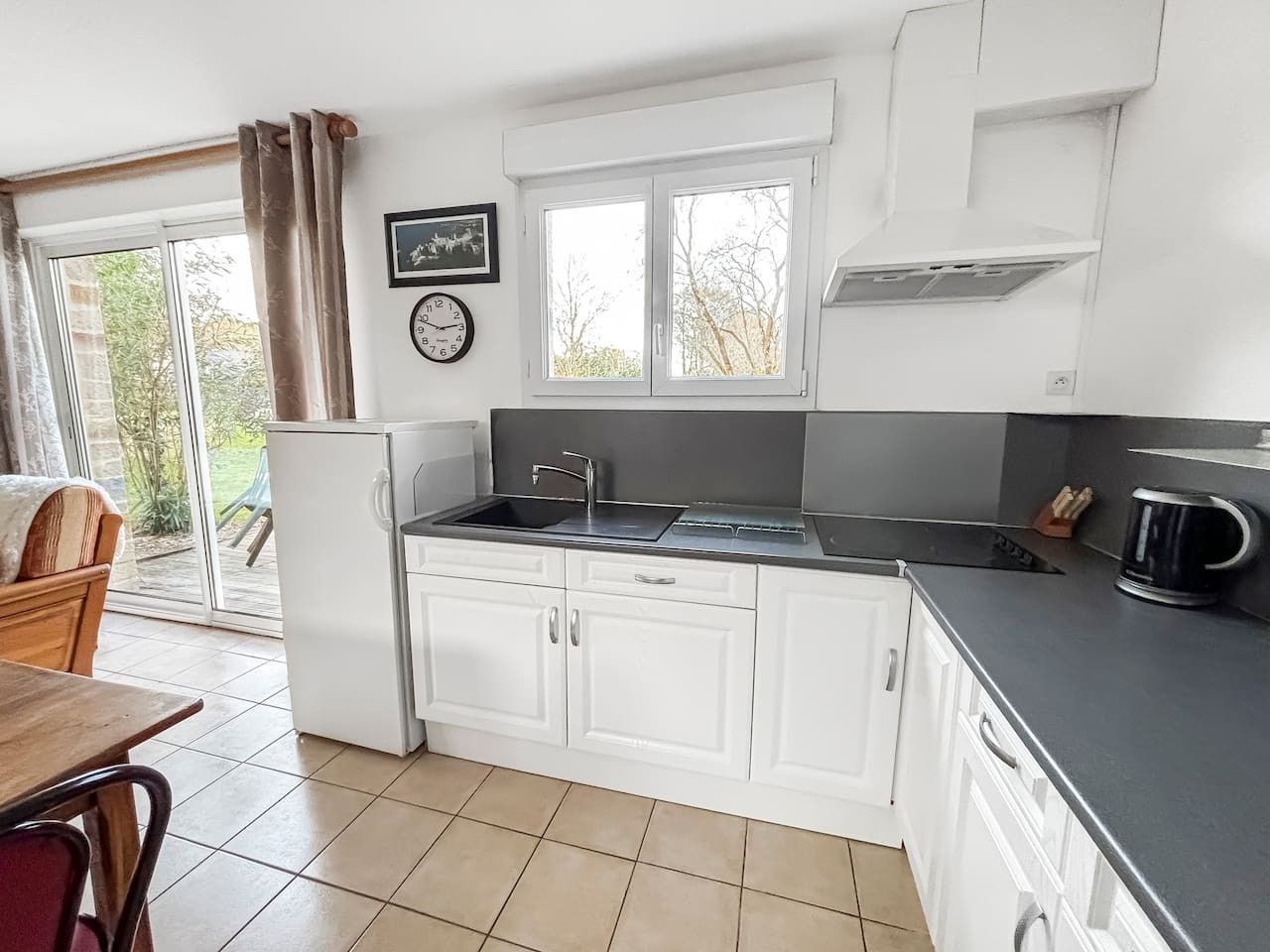 A compact kitchen with white cabinets, a sink, and a stove. There is a window with curtains, a clock, and a picture on the wall.