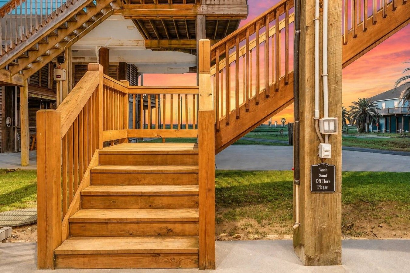 A wooden staircase with a handrail leading to a covered area, a sign instructing to wash sand off here, and a sunset in the background.