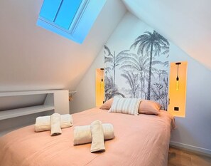 A bedroom with a sloped ceiling, a bed with rolled towels, a wall-mounted light fixture, and a tropical-themed mural. - Cocon Pierre & Lumière, Cœur de Saint-Patern, Vannes (Vannes)