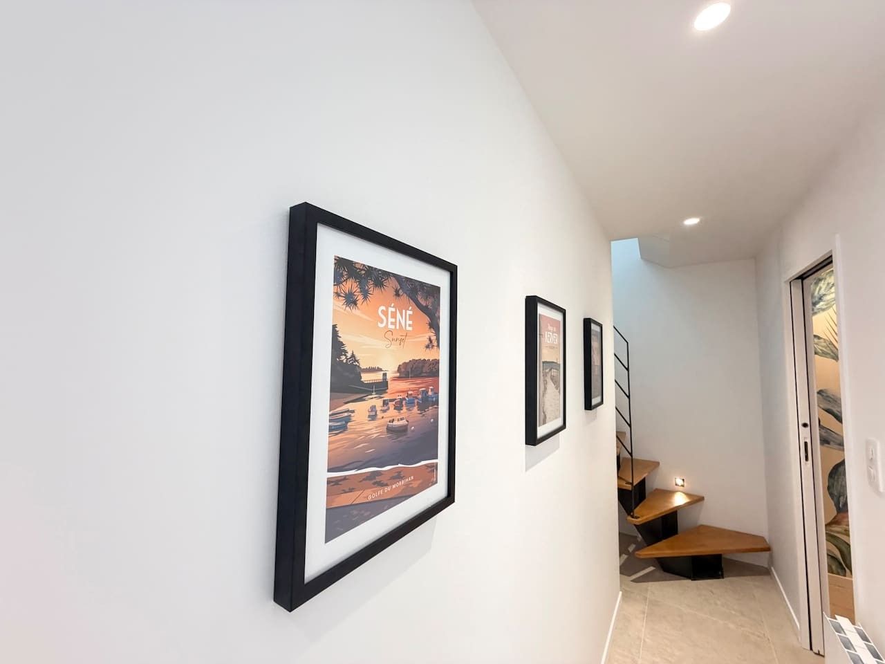 A hallway with framed artwork on the wall, a staircase with wooden steps, and a door leading to another room.