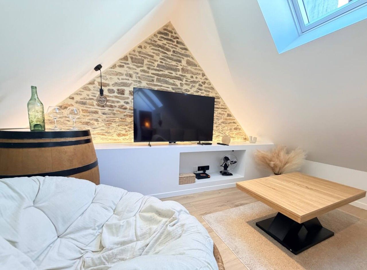 A modern bedroom with a sloped ceiling, a flat-screen TV mounted on a stone wall, a bed with a white duvet, a wooden side table, and a small wooden coffee table.