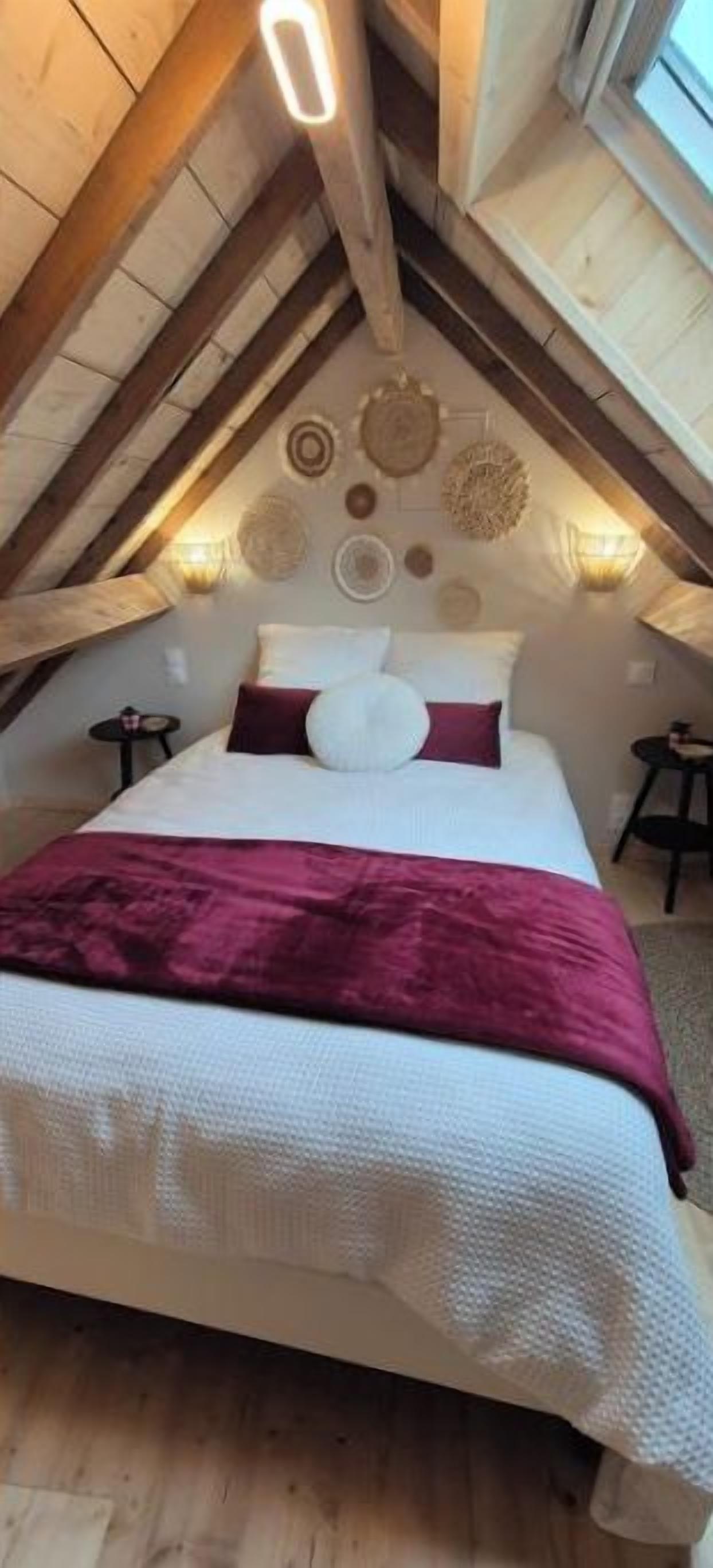 An attic bedroom with a wooden ceiling, a large bed with white bedding and a red throw, and wall-mounted lamps.