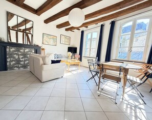 A spacious living area with a sofa, a wooden dining table, and a view of the outside through large windows. - 2-Bedroom Apartment with Exposed Beams – Historic Center of Vannes (Vannes)