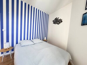 A bedroom with a bed, a bedside table, a lamp, and a wall with vertical stripes. - 2-Bedroom Apartment with Exposed Beams – Historic Center of Vannes (Vannes)