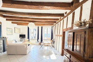 Una habitación con vigas de madera a la vista, suelo de baldosas blancas, un sofá, una mesa de comedor con sillas y un armario.