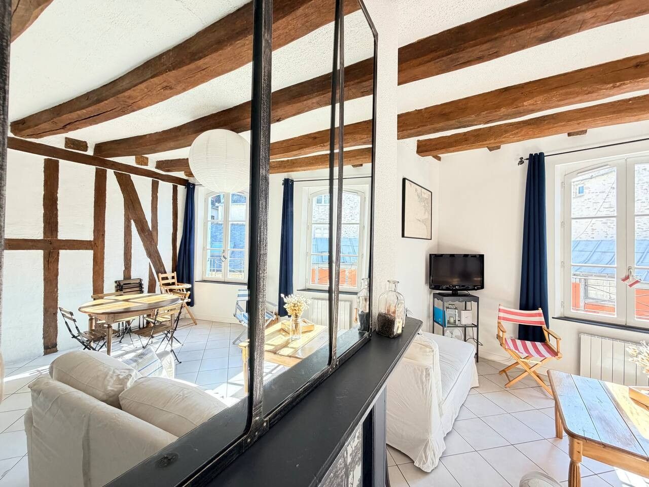 A spacious living area with a high ceiling, wooden beams, a large mirror, a sofa, a dining table, and a television.