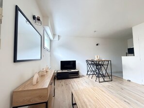 A modern living room with a wooden floor, a flat-screen TV on a black stand, a dining table with chairs, and a large mirror on the wall. - Modern studio apartment in Vannes, just a stone's throw from the city walls (Vannes)